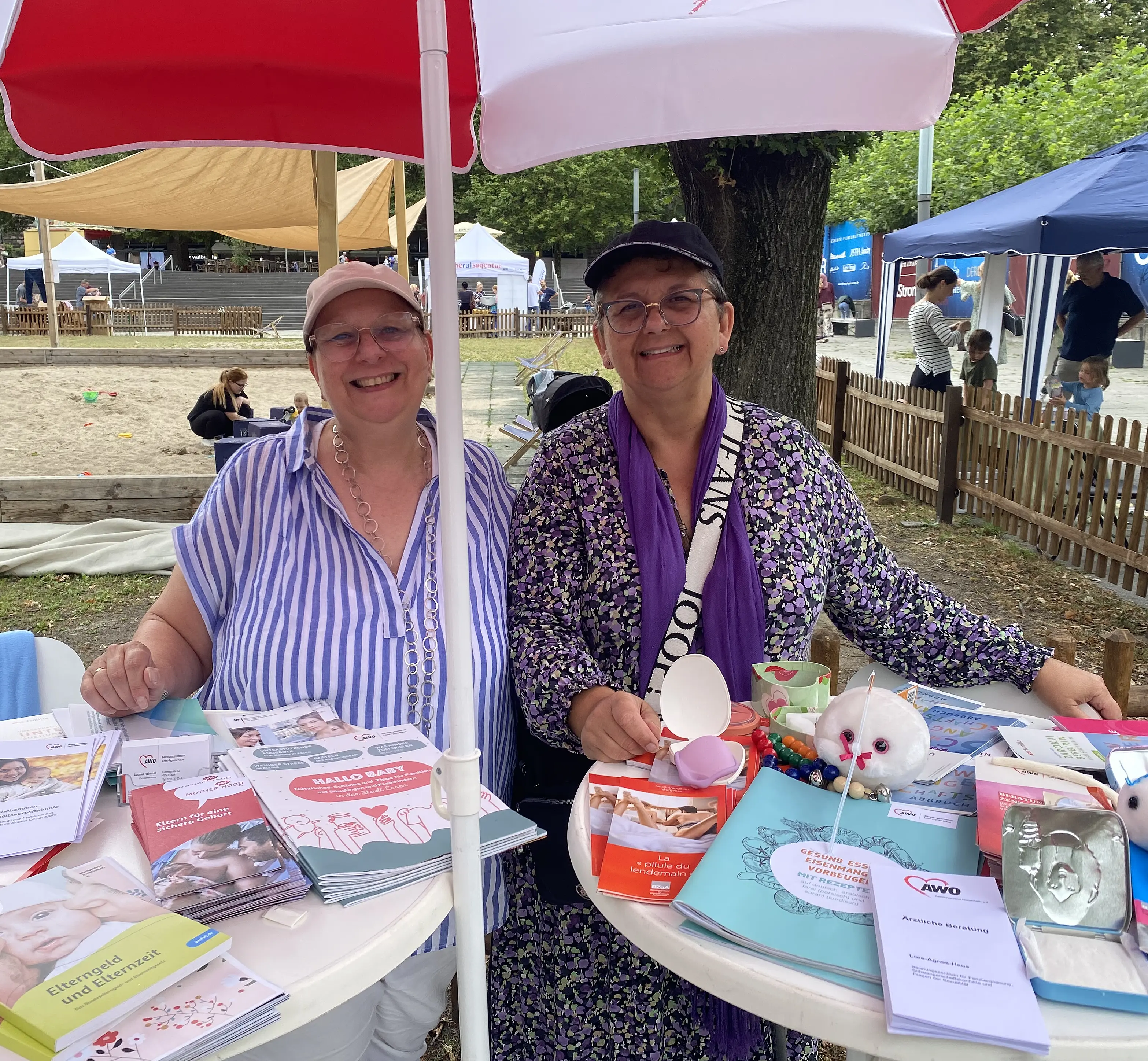 Nahaufnahme von Dagmar Reinhold und Stefanie Boos vor einem Stehtisch mit AWO-Materialien vor dem noch recht leeren Sandkasten.