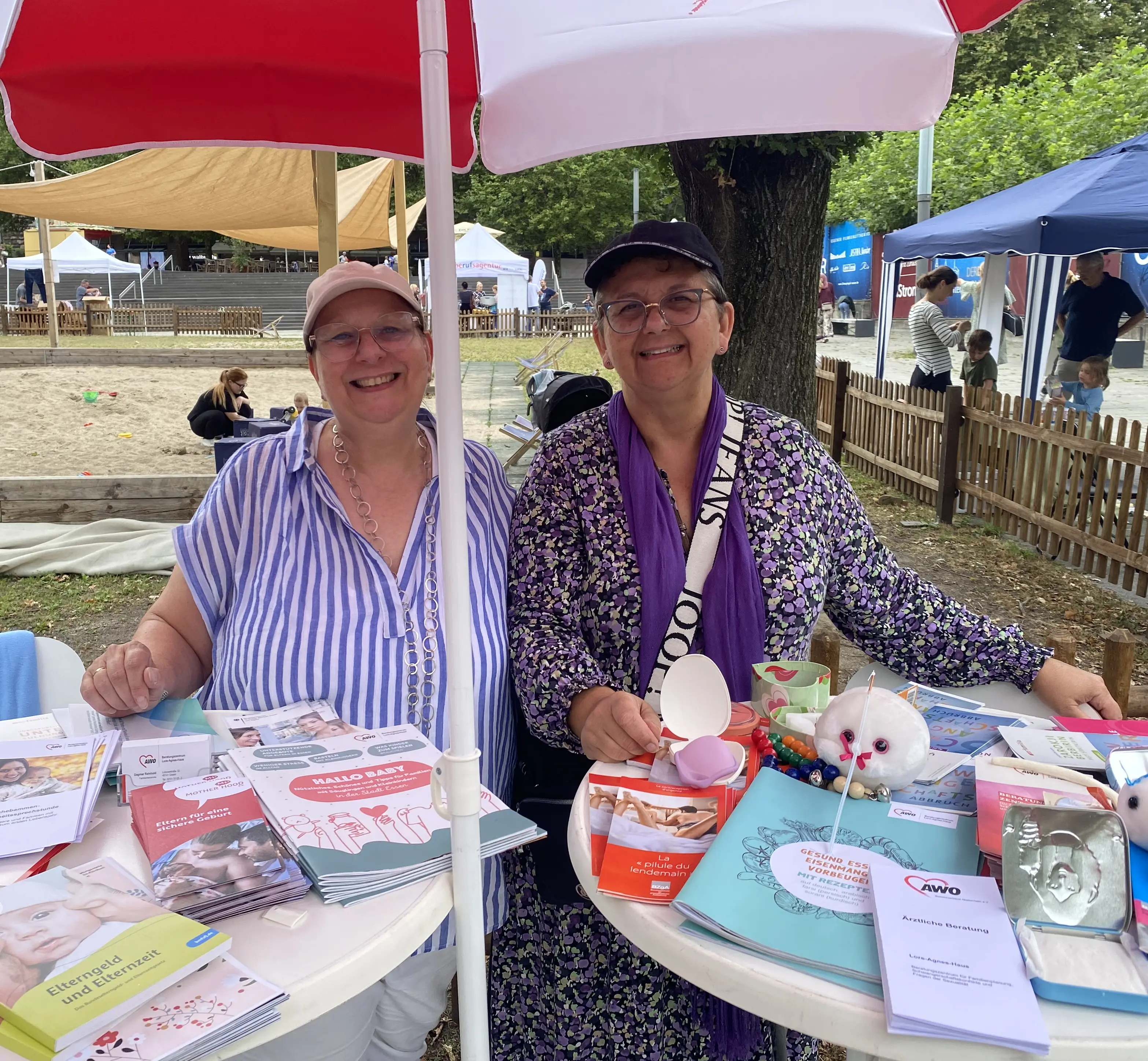 Nahaufnahme von Dagmar Reinhold und Stefanie Boos vor einem Stehtisch mit AWO-Materialien vor dem noch recht leeren Sandkasten.
